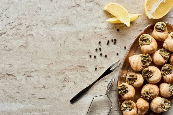 Top view of delicious cooked escargots with lemon slices, black peppercorn and tweezers on stone background — Stock Photo