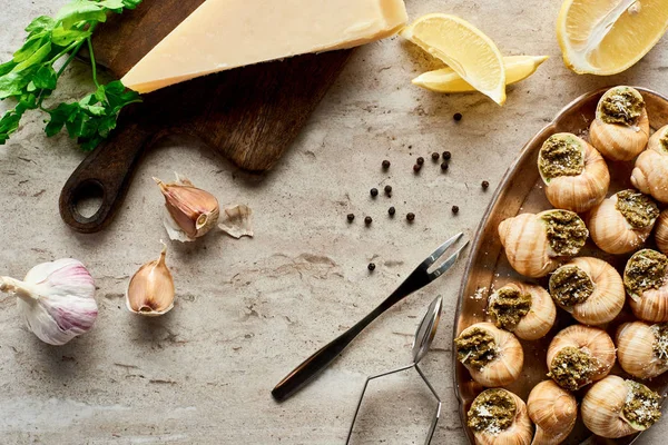 Vista superior de deliciosas escargots cocidas con rodajas de limón, parmesano, ajo, perejil, pimienta negra y pinzas sobre fondo de piedra - foto de stock
