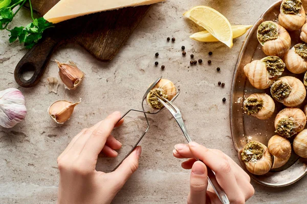 Vista cortada de mulher comer escargots com pinças perto de fatias de limão, parmesão, alho, salsa, pimenta preta no fundo de pedra — Fotografia de Stock