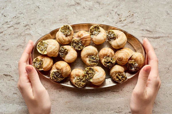 Partial view of woman holding plate with delicious cooked escargots on stone background — Stock Photo