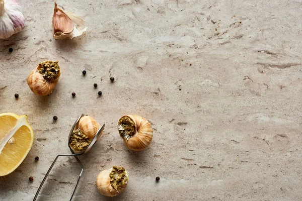 Top view of delicious cooked escargots with lemon, garlic, black peppercorn and tweezers on stone background — Stock Photo