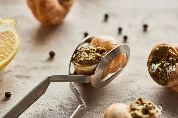 Nahaufnahme köstlicher gekochter Escargots mit Zitrone, schwarzem Pfeffer und Pinzette auf Steinhintergrund — Stockfoto