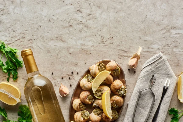 Vista superior de las deliciosas escargots servidas cerca de la botella de vino blanco y los ingredientes sobre fondo de piedra - foto de stock