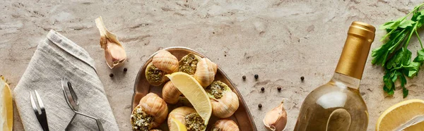 Vista superior de las deliciosas escargots servidas cerca de la botella de vino blanco y los ingredientes sobre fondo de piedra, plano panorámico - foto de stock