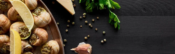 Vista superior de deliciosas escargots cocidas con rodajas de limón en mesa de madera negra con ingredientes, plano panorámico - foto de stock