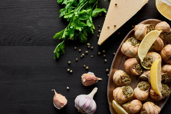 Top view of delicious cooked escargots with lemon slices on black wooden table with ingredients — Stock Photo