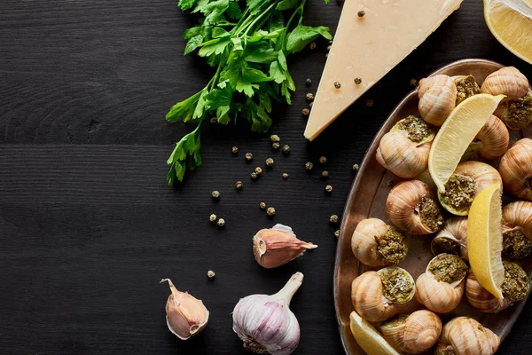 Vista superior de deliciosos escargots cozidos com fatias de limão em mesa de madeira preta com ingredientes — Fotografia de Stock