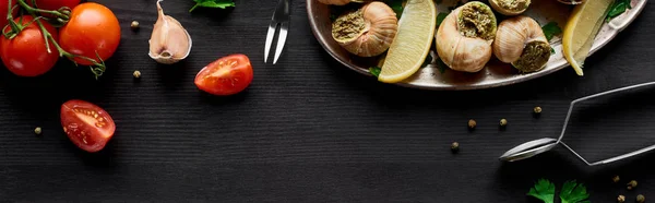 Top view of delicious cooked escargots with lemon slices on black wooden table with ingredients, panoramic shot — Stock Photo