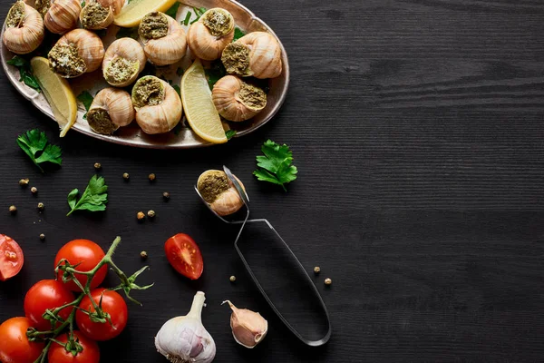 Top view of delicious cooked escargots on black wooden table with ingredients and tweezers — Stock Photo