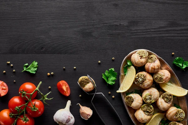 Top view of delicious cooked escargots on black wooden table with ingredients and tweezers — Stock Photo