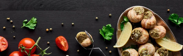 Top view of delicious cooked escargots on black wooden table with ingredients and tweezers, panoramic shot — Stock Photo