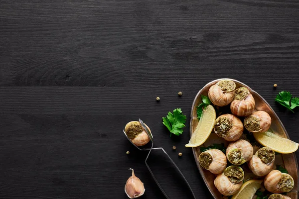 Vista superior de deliciosas escargotas cozidas com limão e pinças em mesa de madeira preta — Fotografia de Stock