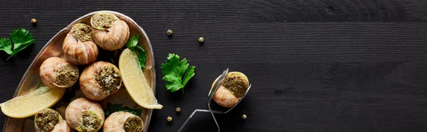 Vista superior de deliciosas escargotas cozidas com limão e pinças em mesa de madeira preta, tiro panorâmico — Fotografia de Stock