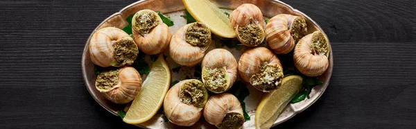 Top view of delicious cooked escargots with lemon on black wooden table, panoramic shot — Stock Photo