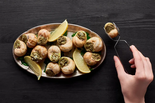 Cropped view of woman eating delicious escargots with lemon on black wooden table — Stock Photo