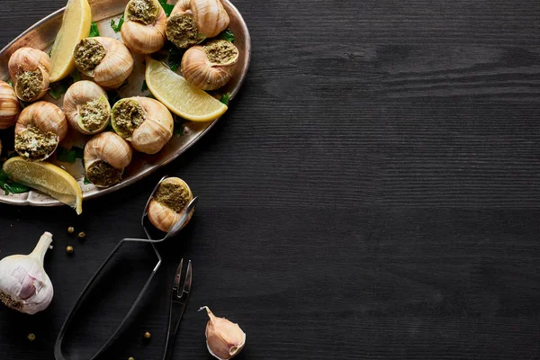 Vista superior de deliciosas escargotas cozidas com limão e pinças em mesa de madeira preta — Fotografia de Stock