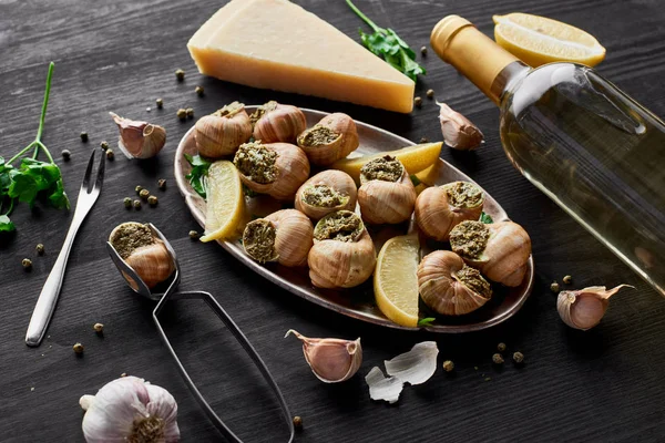 Delicious cooked escargots with lemon slices on black wooden table with spices, Parmesan and white wine — Stock Photo