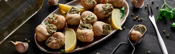 Delicious cooked escargots with lemon slices on black wooden table with spices and white wine, panoramic shot — Stock Photo