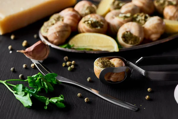 Foco seletivo de deliciosas escargotas cozidas com fatias de limão perto de talheres, pimenta preta e parmesão em mesa de madeira preta — Fotografia de Stock