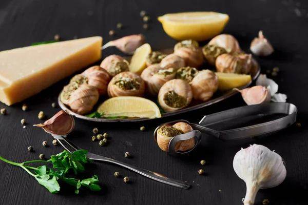 Enfoque selectivo de deliciosas escargots cocidas con rodajas de limón cerca de ajo, cubiertos, pimienta negra y parmesano en mesa de madera negra — Stock Photo