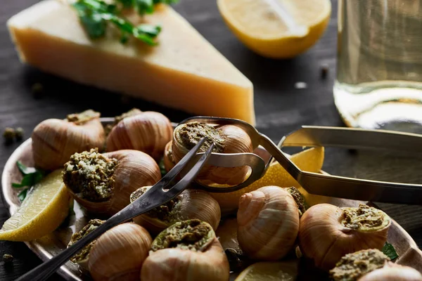 Selective focus of delicious cooked escargots with lemon, parmesan, cutlery, parsley and white wine on black wooden table — Stock Photo