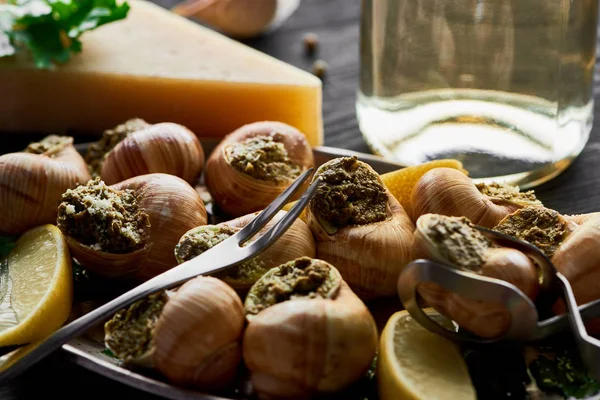 Foco seletivo de deliciosas escargotas cozidas com limão, parmesão, talheres, salsa e vinho branco na mesa de madeira preta — Fotografia de Stock