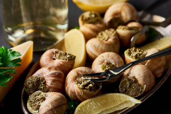Selective focus of delicious cooked escargots with lemon, parmesan, cutlery, parsley and white wine on black wooden table — Stock Photo