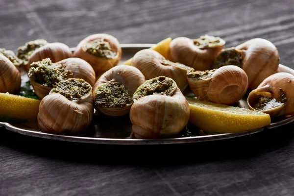 Vista de cerca de deliciosas escargots cocidas con rodajas de limón en el plato en la mesa de madera negro - foto de stock