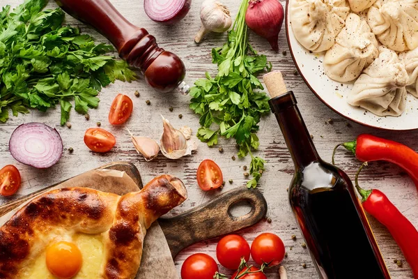 Top view of delicious Adjarian khachapuri and khinkali near bottle of wine, vegetables and spices on wooden table — Stock Photo