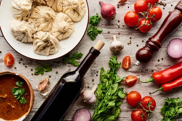 Вид на вкусный хинкали возле бутылки вина, харчо, овощей и специй на деревянном столе — стоковое фото