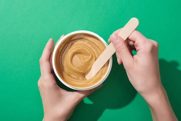 Cropped view of woman holding stick and cup with depilation wax on green background — Stock Photo