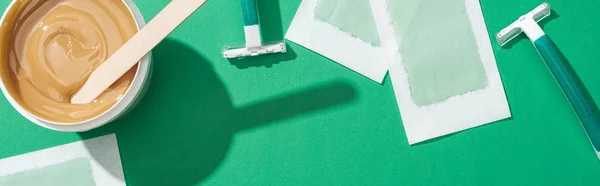 Top view of green disposable razors, wax and depilation stripes on green background, panoramic shot — Stock Photo