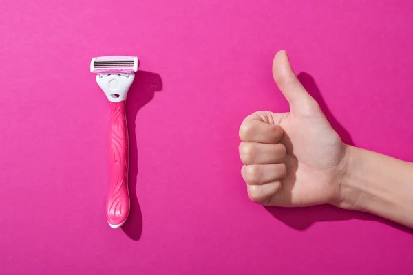 Vista recortada de la mujer que muestra el pulgar hacia arriba cerca de la navaja de afeitar sobre fondo rosa - foto de stock