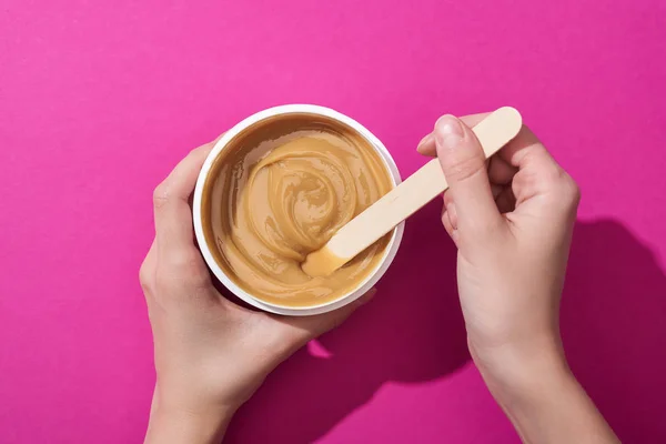 Cropped view of woman holding cup with depilation wax and stick on pink background — Stock Photo