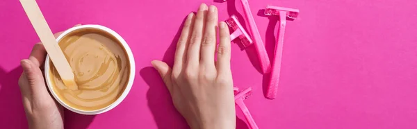 Vista recortada de la mujer alejándose afeitadoras desechables y tomando depilación cera en taza con palo sobre fondo rosa, tiro panorámico - foto de stock