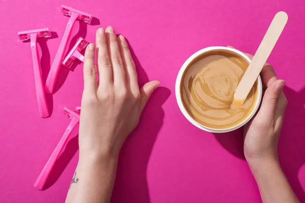 Cropped view of woman moving away disposable razors and taking depilation wax in cup with stick on pink background — Stock Photo