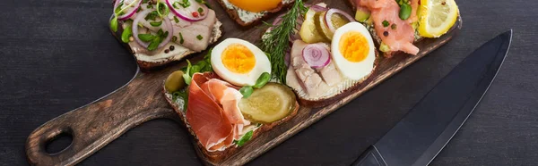 Panoramic shot of wooden cutting board with danish smorrebrod sandwiches near knife on grey surface — Stock Photo