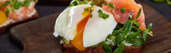 Panoramic shot of danish smorrebrod sandwich with poached egg near salmon on wooden cutting board — Stock Photo