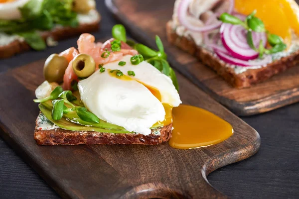 Close up de saboroso sanduíche de smorrebrod com ovo escalfado perto de salmão fresco em tábua de corte de madeira — Fotografia de Stock