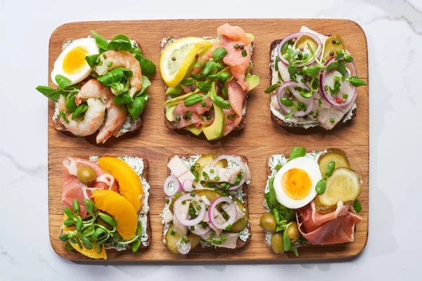 Vue du dessus de la planche à découper en bois avec de savoureux sandwichs smorrebrod sur la surface en marbre blanc — Photo de stock