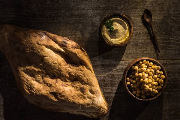 Blick von oben auf köstlichen Hummus und Kichererbsen in Schüsseln in der Nähe von frisch gebackener Pita und Löffel auf rustikalem Holztisch — Stockfoto