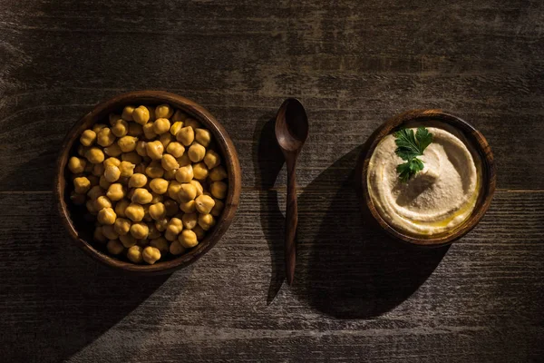 Vista superior de delicioso hummus e grão de bico em tigelas perto de colher em mesa rústica de madeira — Fotografia de Stock