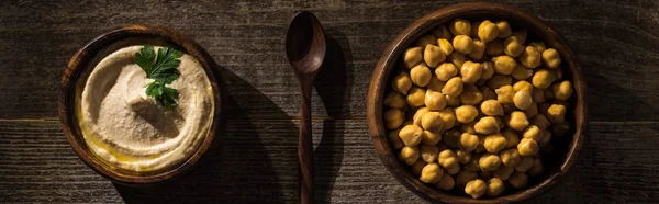 Vue de dessus de délicieux houmous et pois chiches dans des bols près de cuillère sur table rustique en bois, vue panoramique — Photo de stock
