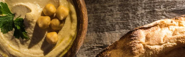 Vista superior de delicioso hummus com grão de bico em tigela perto de pita cozido na mesa rústica de madeira, tiro panorâmico — Fotografia de Stock