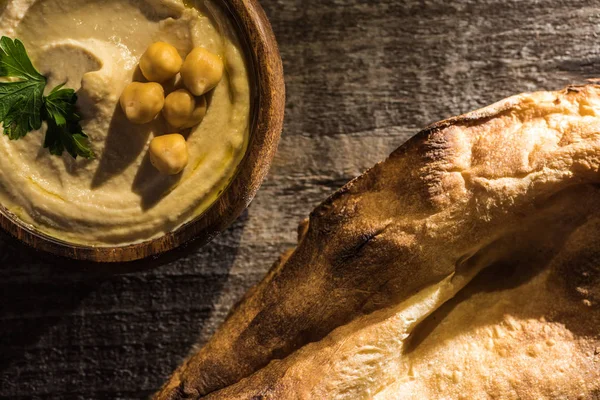 Vista superior de delicioso hummus com grão de bico na tigela perto de pita cozido na mesa rústica de madeira — Fotografia de Stock