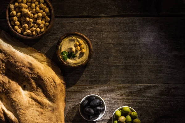 Vista superior de delicioso hummus, garbanzos, pita y aceitunas en la mesa rústica de madera - foto de stock