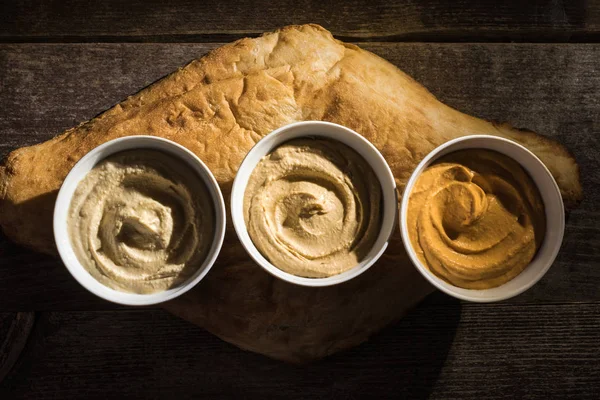 Draufsicht auf köstlich sortierten Humus auf frisch gebackenem Fladenbrot auf rustikalem Holztisch — Stockfoto