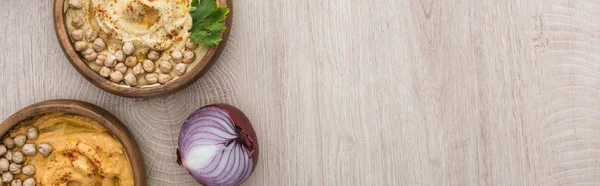 Vue du dessus de délicieux houmous aux pois chiches dans un bol près de l'oignon rouge sur une table en bois beige, vue panoramique — Photo de stock