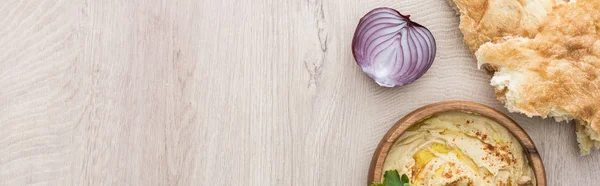 Top view of delicious hummus in bowl near fresh baked pita, red onion on beige wooden table, panoramic shot — Stock Photo