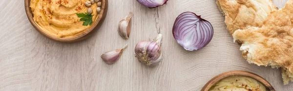 Vista dall'alto di delizioso hummus in ciotola vicino alla pita appena sfornata, aglio e cipolla rossa sul tavolo di legno beige, colpo panoramico — Foto stock